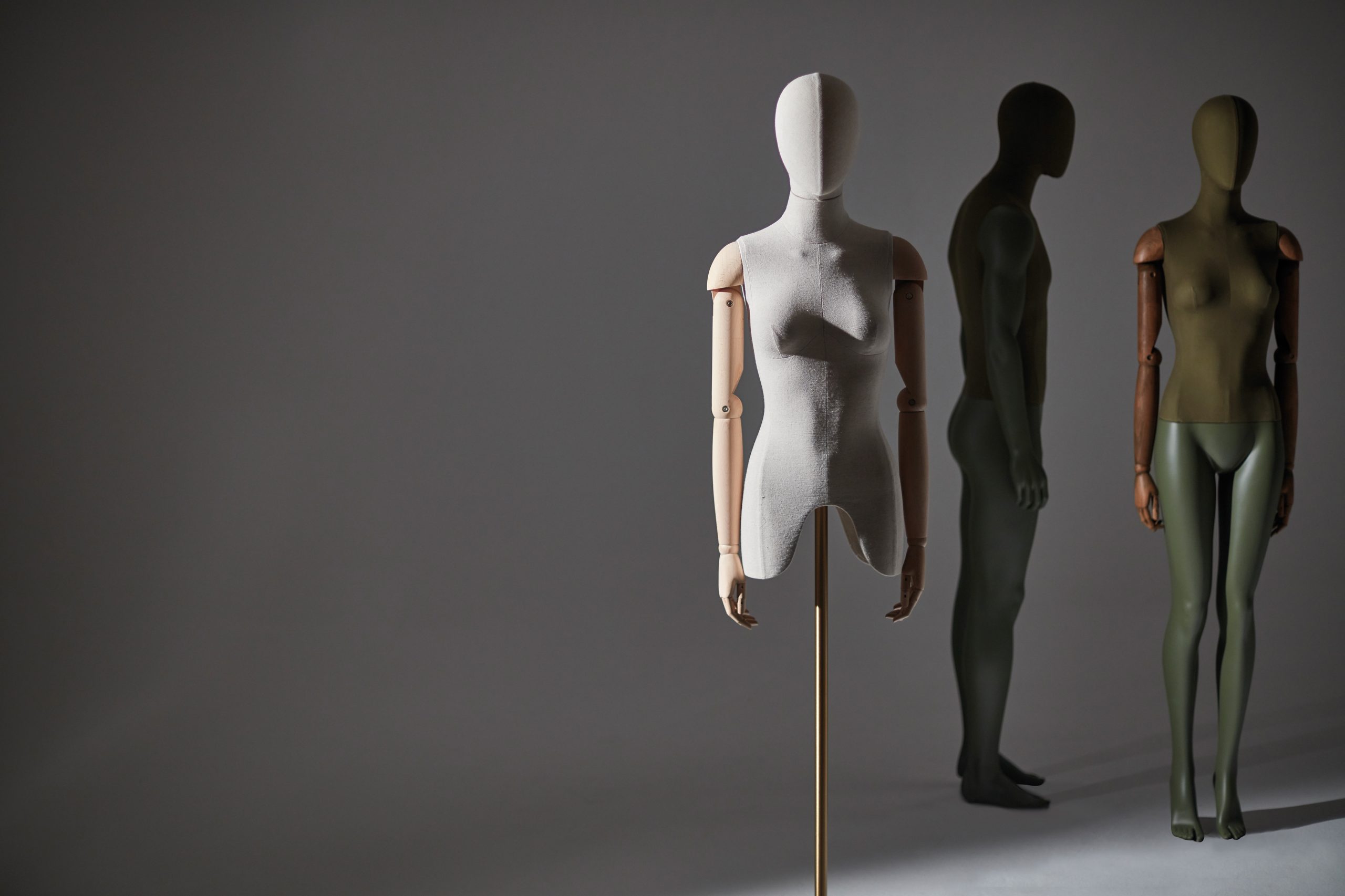 Mannequin by HOL Three mannequins in a dimly lit space. The mannequin in the foreground is light-colored, headless, armless, and stands on a pole base. Behind it, two bespoke full-body mannequins, one light and the other dark, face each other, partially shadowed.