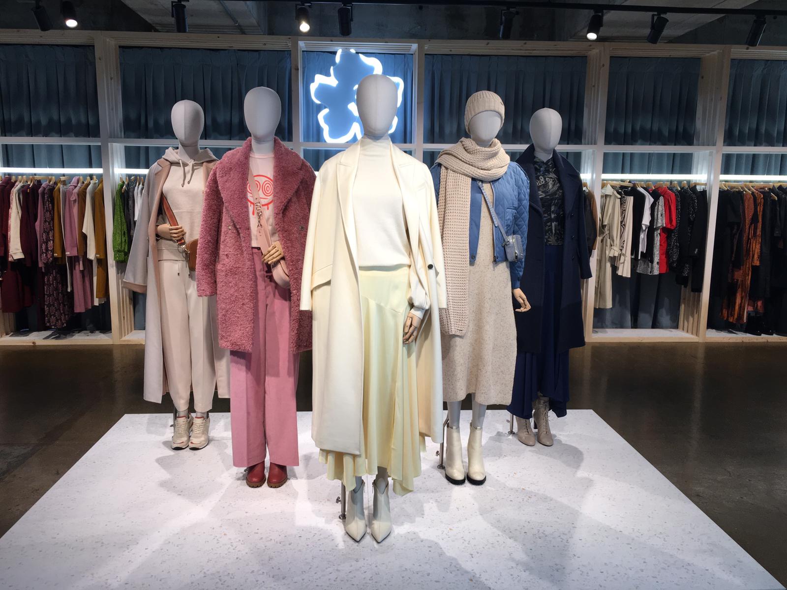 Mannequin by HOL A display of five mannequins dressed in various stylish, personalized outfits stands in front of a rack of clothes. The outfits range from casual to elegant, featuring long coats, sweaters, and layered garments in shades of pink, cream, beige, and denim.