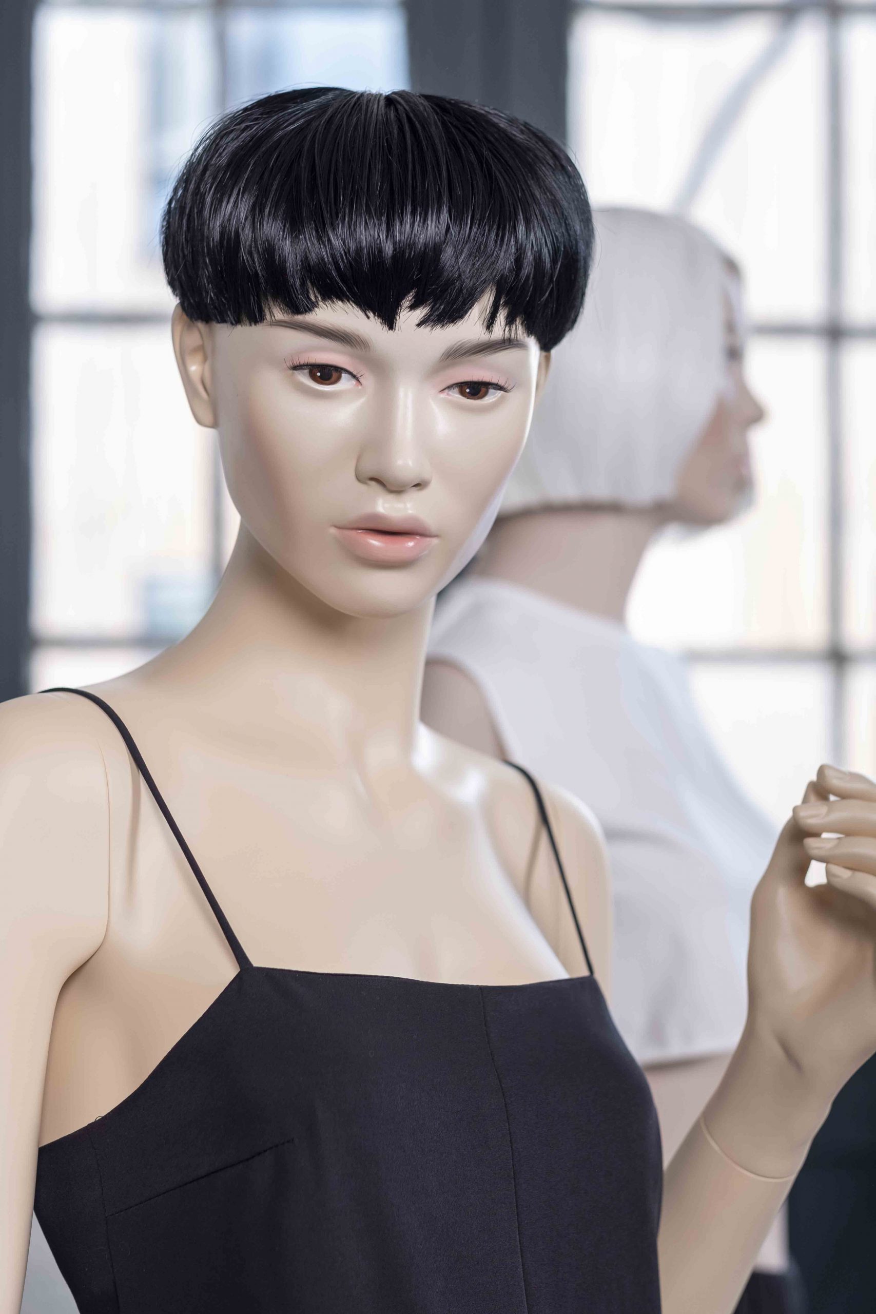 Mannequin by HOL Two mannequins are displayed in a brightly lit room with large windows. The foreground mannequin features a short, black bob haircut and is dressed in a sleeveless black outfit with thin straps, showcasing custom tailoring. The background mannequin has a short, white haircut and wears a simple white top.