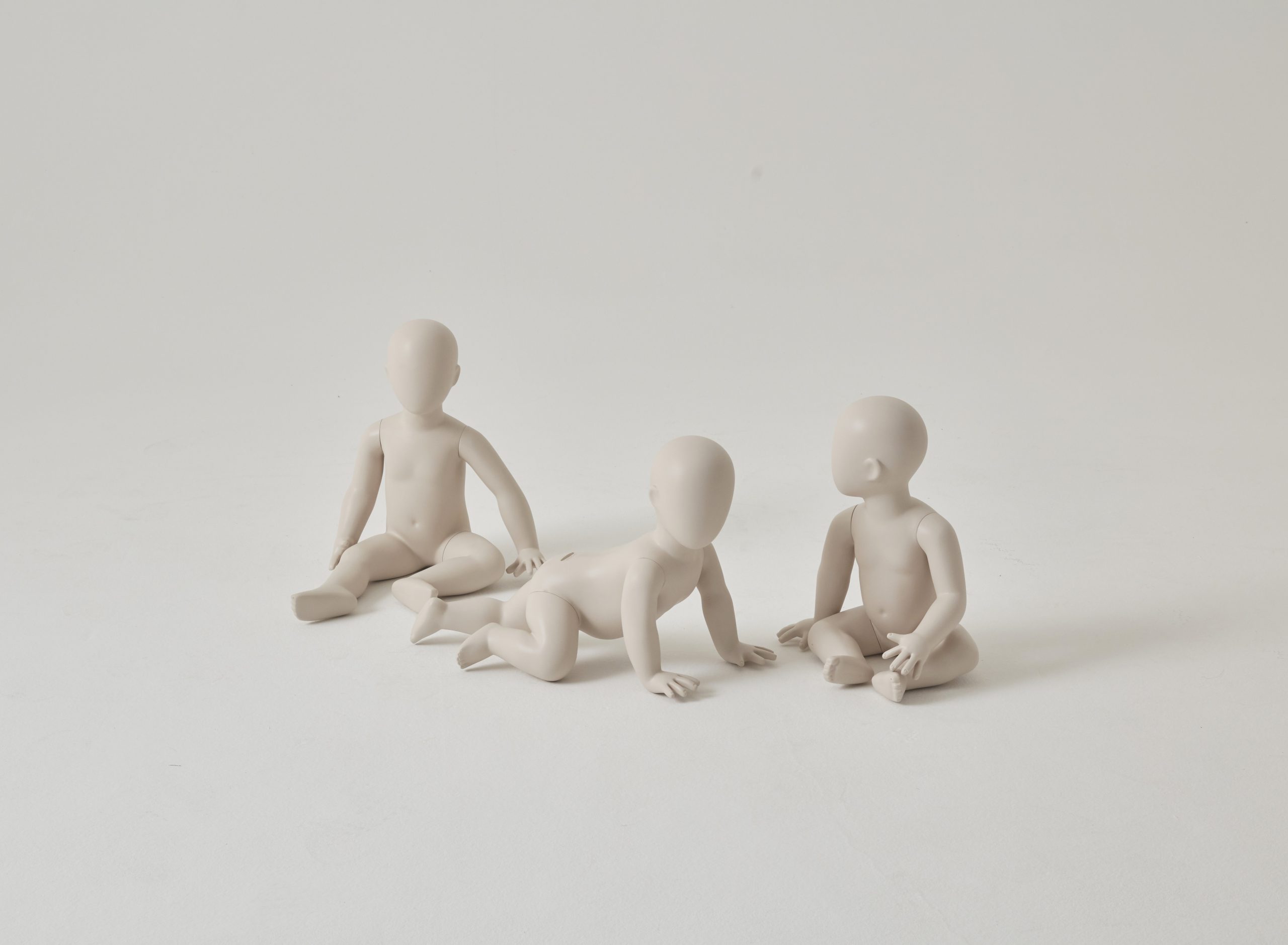 Mannequin by HOL Three simple, beige, faceless human-like figurines resemble child mannequins seated on a white background. One figure has legs stretched out, another is positioned on all fours, and the third is sitting cross-legged. All convey a childlike form.