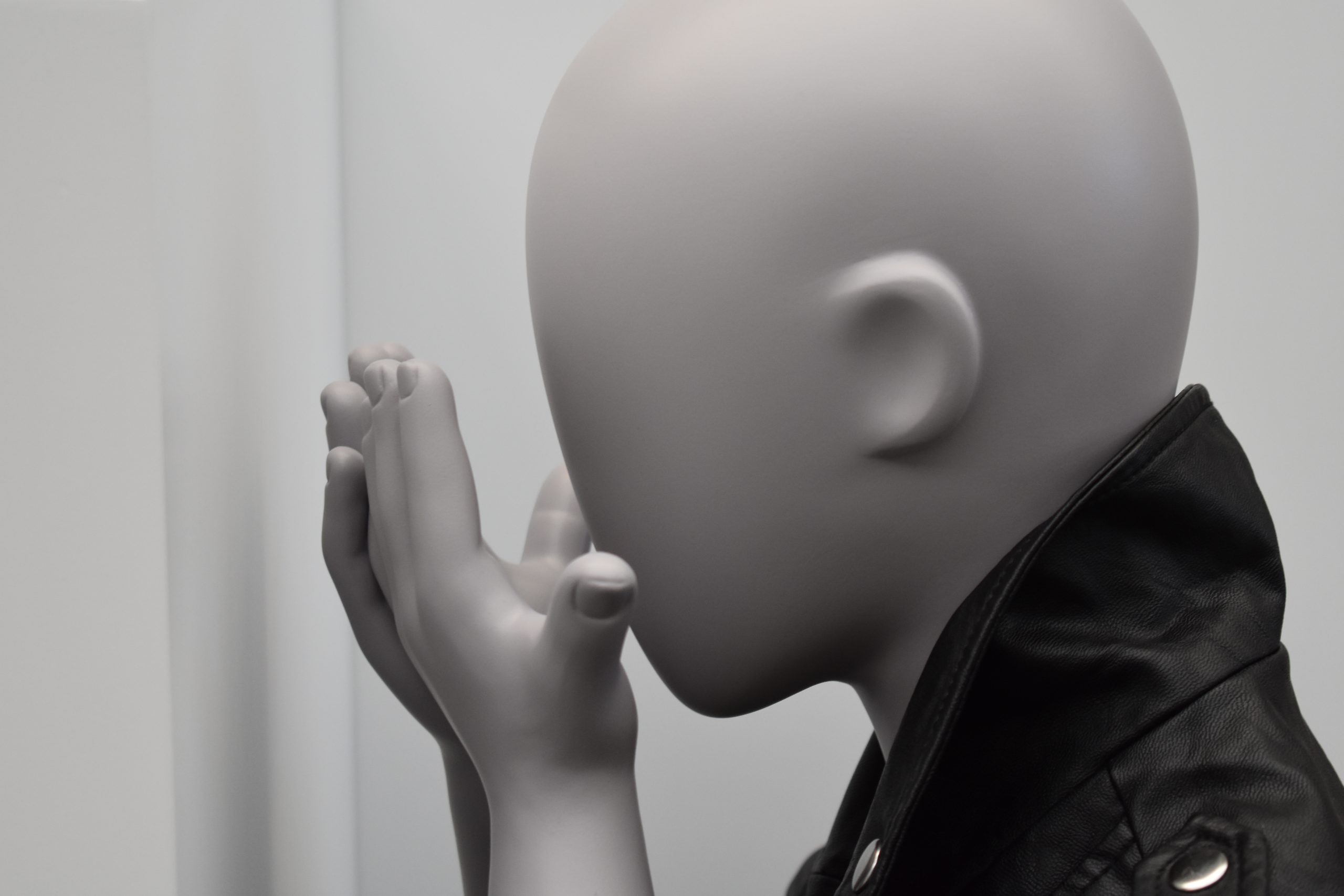 Mannequin by HOL A faceless child mannequin with smooth, featureless skin is posed with its hands gently pressed together near its mouth. The mannequin is wearing a black leather jacket and is set against a plain, light-colored background.