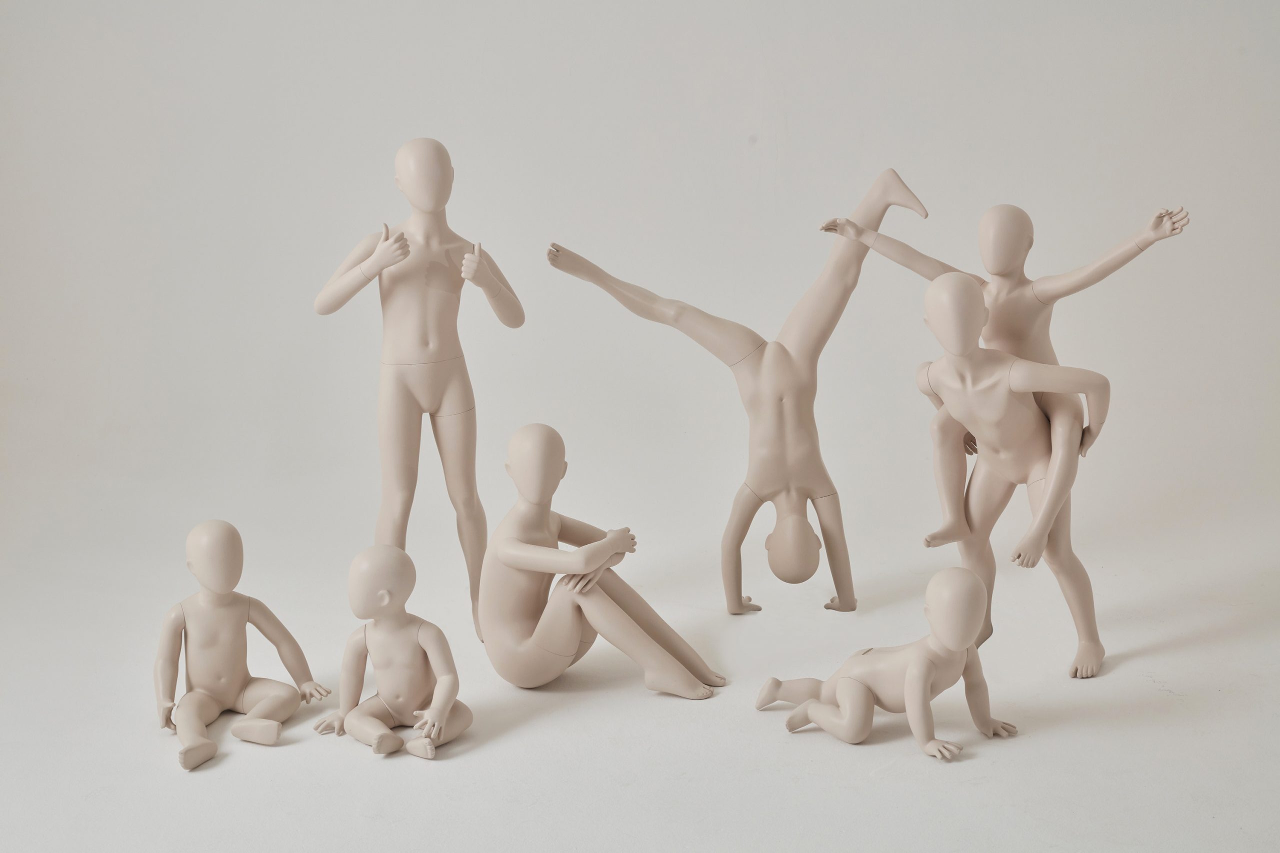 Mannequin by HOL A group of minimalistic, flesh-colored human figurines, resembling child mannequins, is arranged in various poses. Some are seated, while others are standing, doing handstands, or piggybacking. The background is plain white, making the figures the focal point.