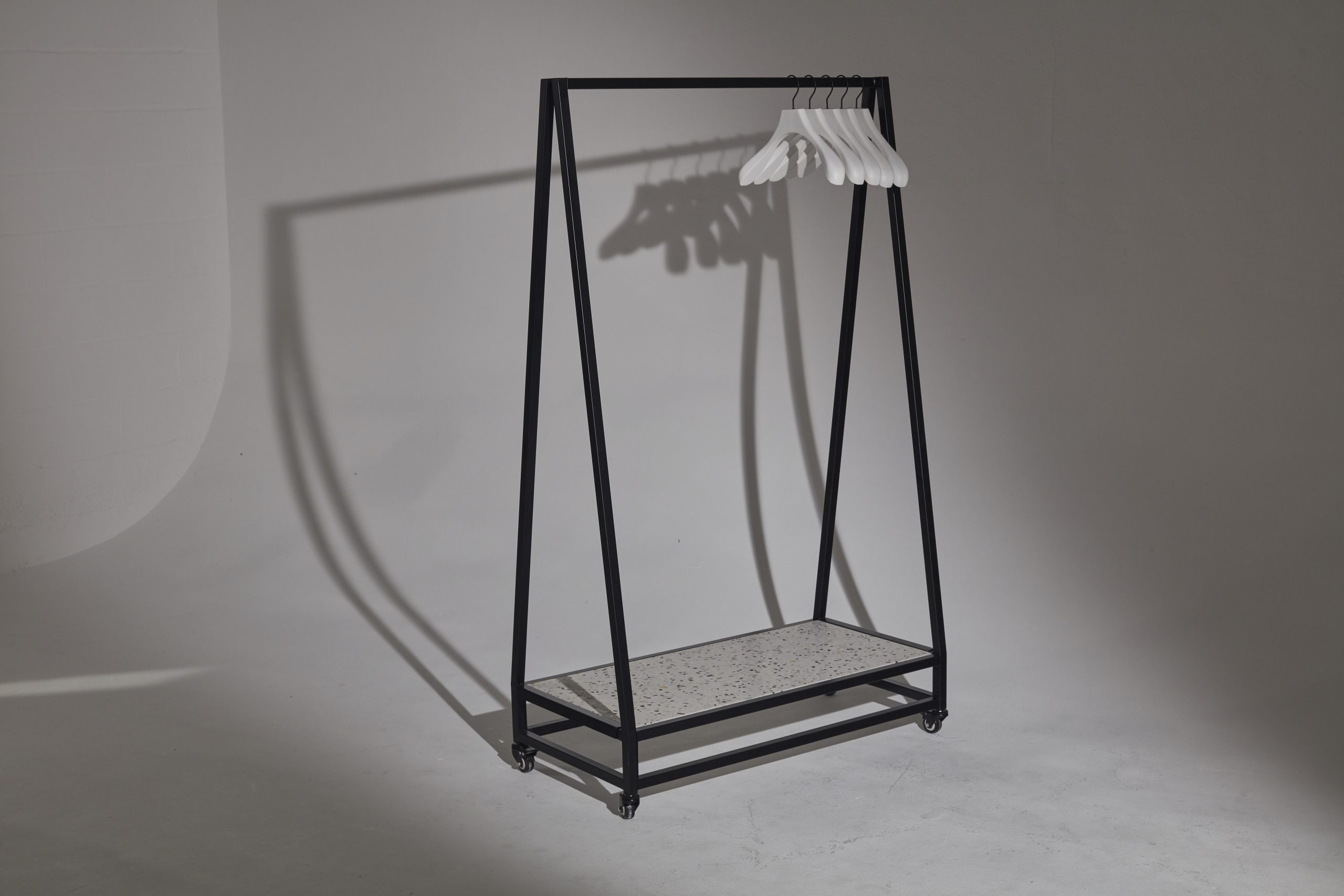 Mannequin by HOL A minimalist black clothing rack with a triangular frame stands in an empty, well-lit room. The piece of furniture features six white hangers dangling from it and a speckled lower shelf. The plain background casts shadows from the rack and hangers onto the floor and wall. Perfect for hire as stylish props in photoshoots.