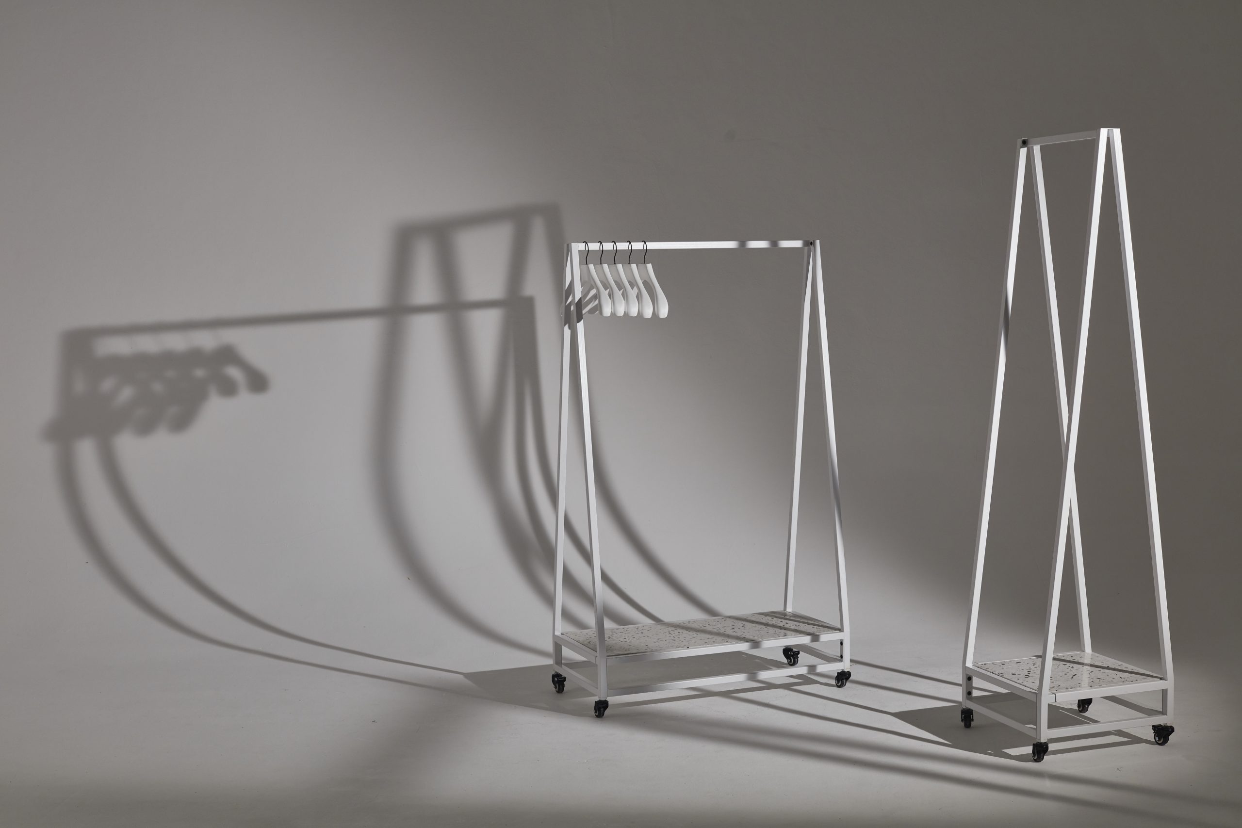 Mannequin by HOL A minimalist metal clothes rack on wheels with a few empty white hangers is positioned next to a taller, narrow triangular metal stand. Both cast shadows on a plain gray background. The setup is simple and modern, highlighting geometric shapes and clean lines—ideal for hire as stylish furniture props.