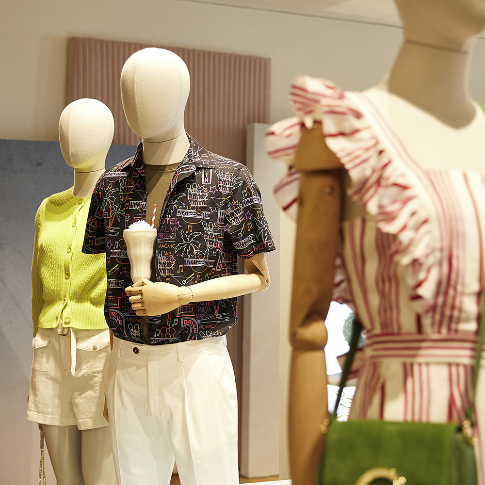 Mannequin by HOL Three mannequins are dressed in summer outfits. The left mannequin wears a green top and white shorts. The center mannequin is in a patterned shirt and white pants, holding a white item. The right mannequin sports a pink and white striped dress with ruffled sleeves and a green purse.