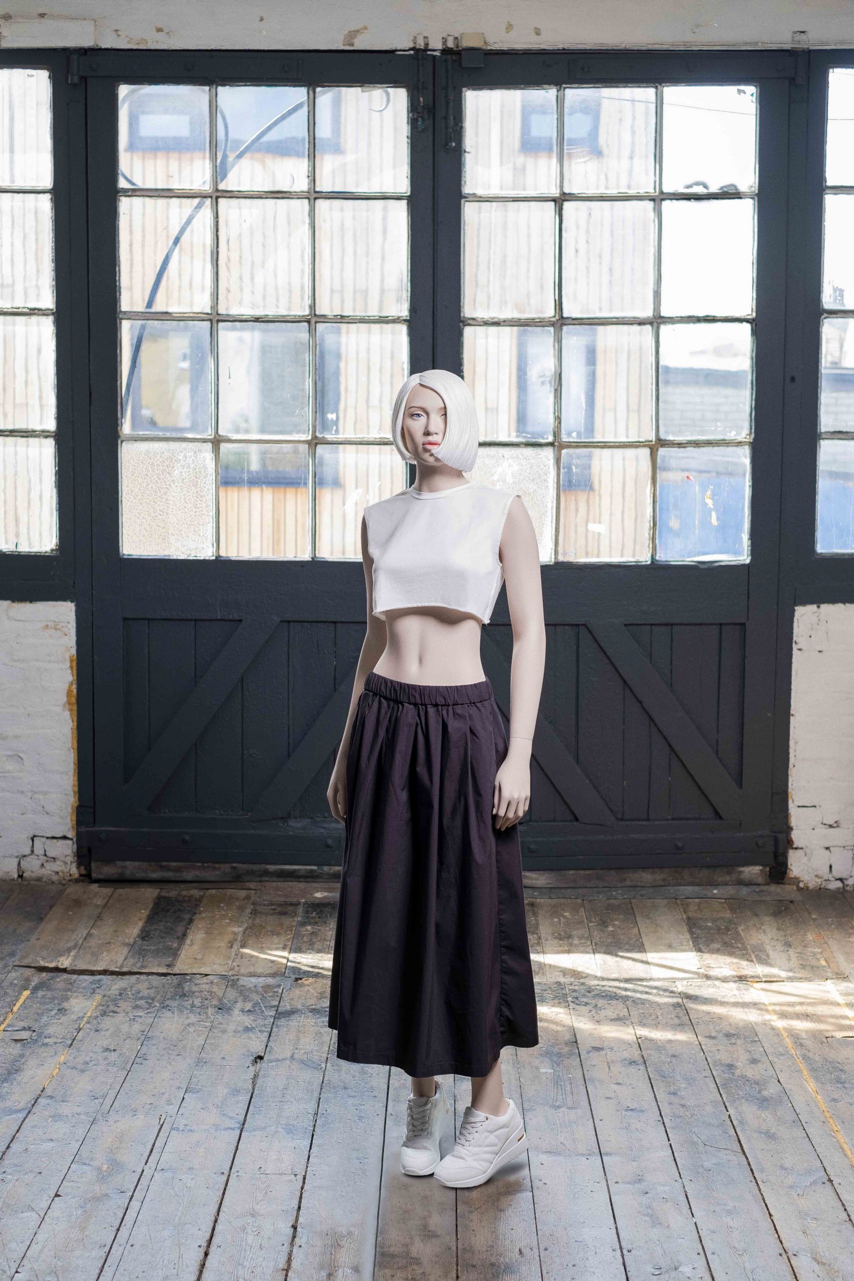 Mannequin by HOL A mannequin with a white, sleeveless crop top and a long, black skirt stands on a wooden floor. The scene is set in front of large industrial-style windows with a mix of brick and wooden walls. The mannequin, perfect for Look Books, is also wearing white sneakers.