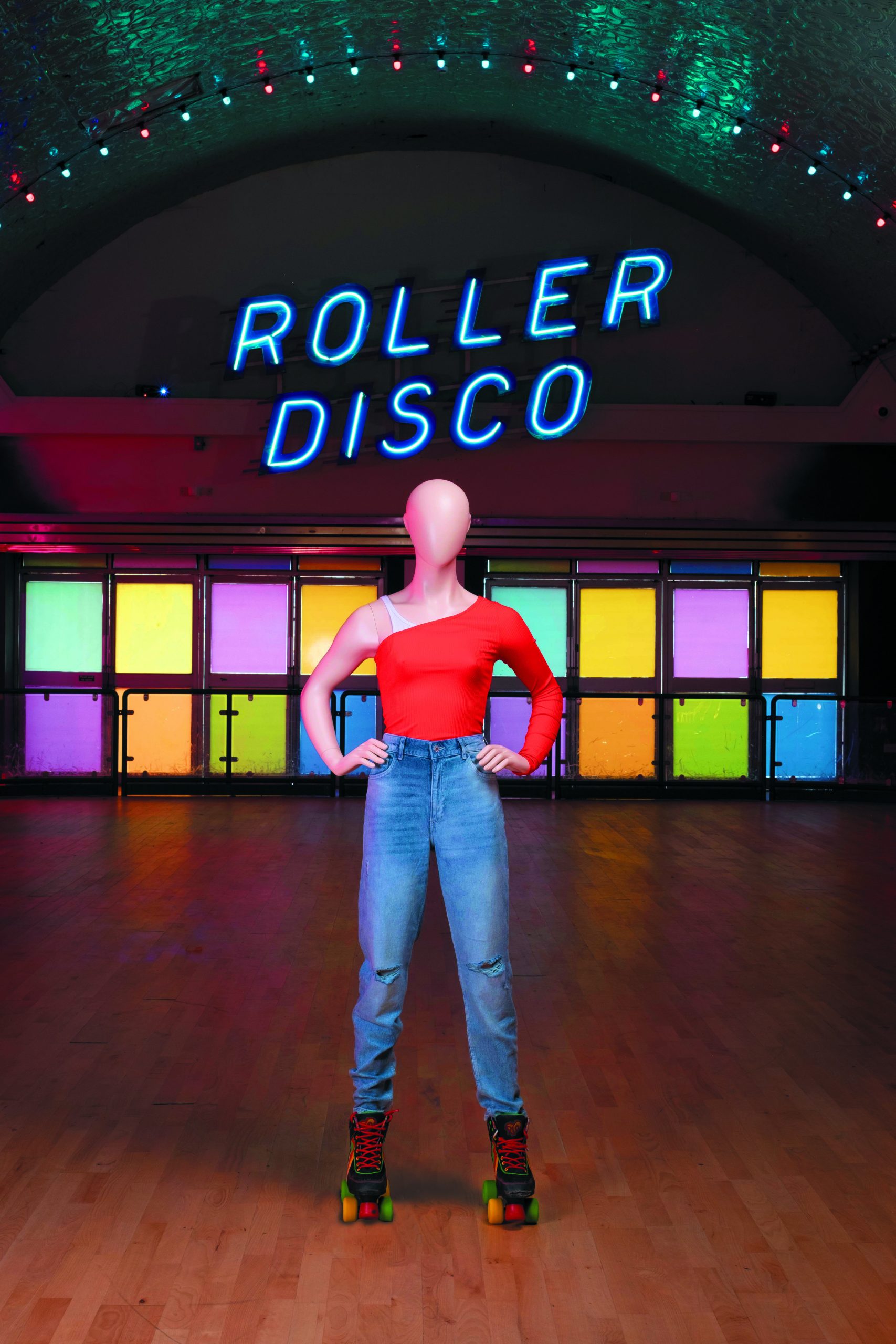 Mannequin by HOL A mannequin in a red one-shoulder top, ripped jeans, and roller skates stands on a roller rink floor beneath a glowing neon "Roller Disco" sign. The scene, perfect for Look Books, features colorful, illuminated panels in the background.