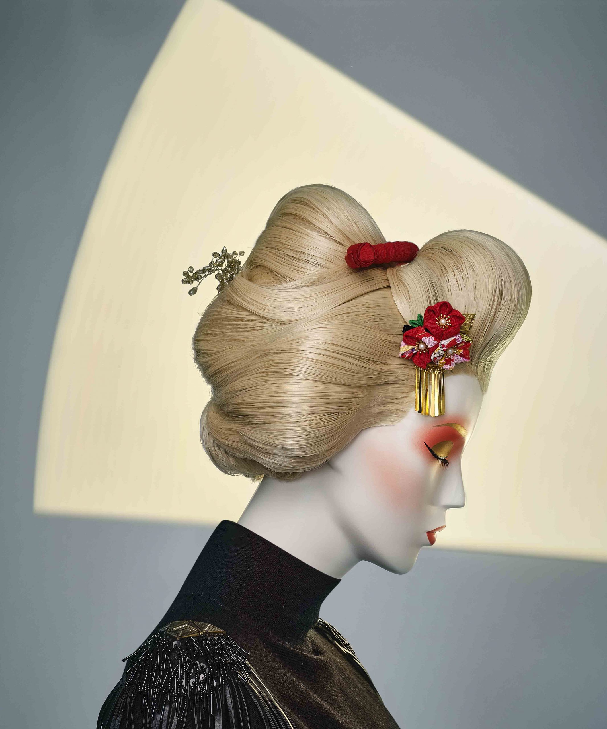 Mannequin by HOL A person with elaborate blonde hair adorned with traditional Japanese ornaments, including red flowers and golden hairpins, is shown in profile. They wear black attire and dramatic makeup against a muted background with a triangular, light-colored shape, reminiscent of high-fashion Look Books.