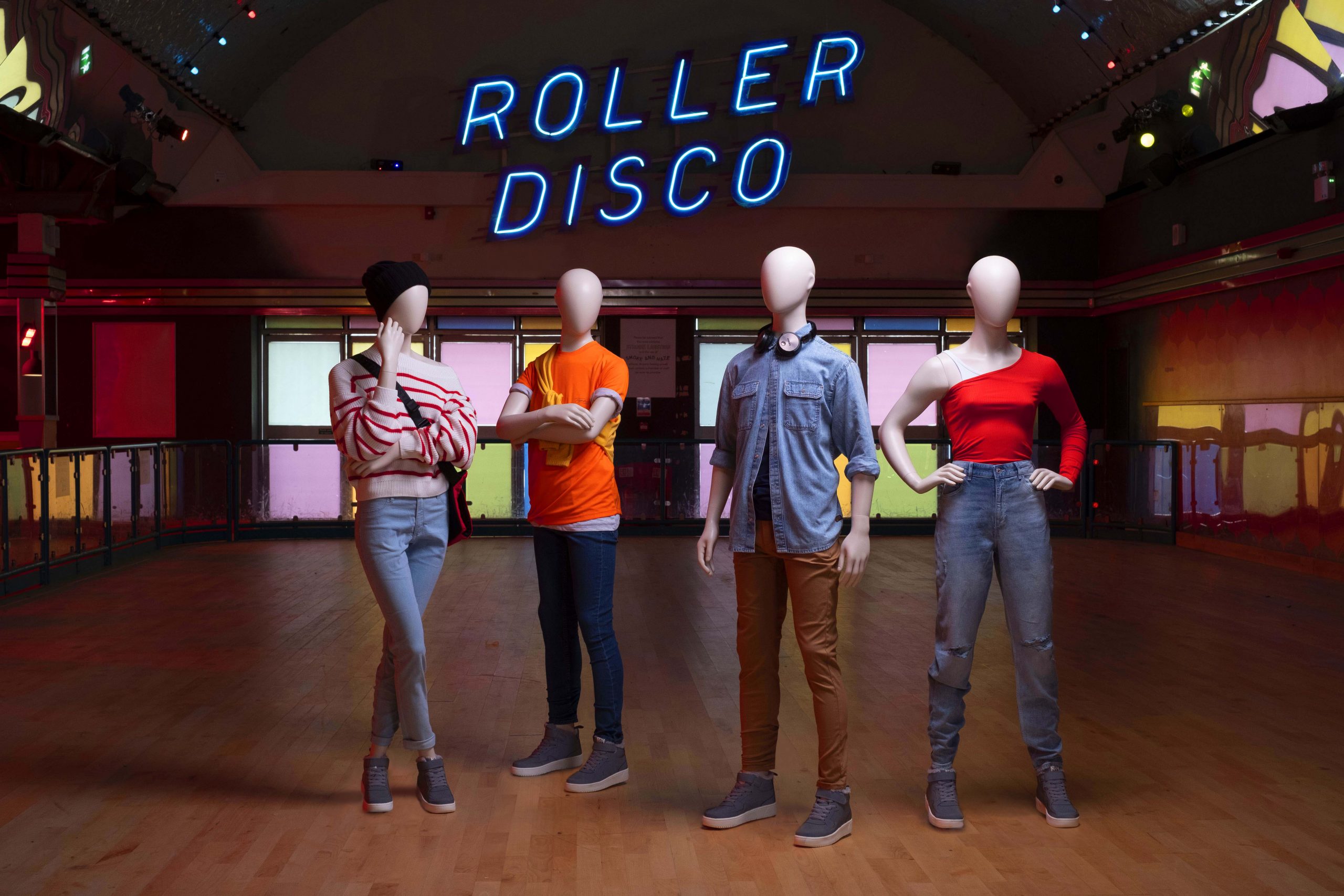 Mannequin by HOL Four mannequins in casual attire stand in Farringdon Youth's retro-styled roller disco with neon lights above them spelling "Roller Disco." The setting features a wooden floor and a large arched ceiling with additional colorful lighting, perfect for youth programs.