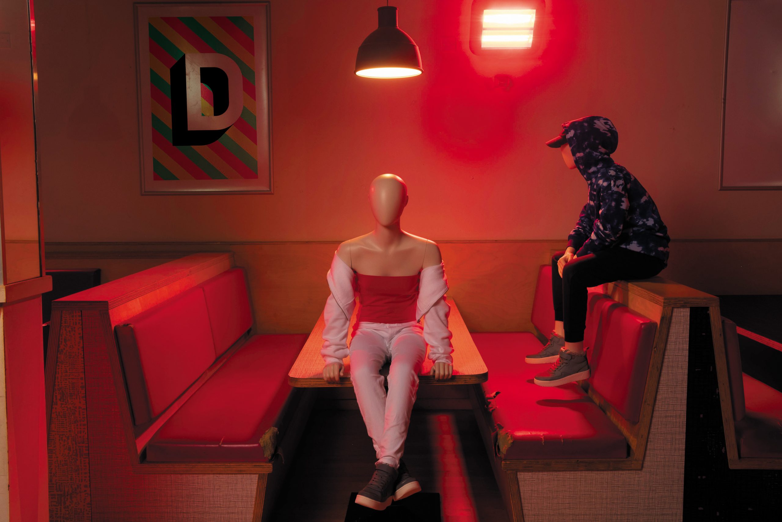 Mannequin by HOL A dimly lit diner with red booths. A mannequin dressed in a red and white outfit sits at a table under a hanging light. A person in a hoodie and cap, possibly one of the Farringdon Youth, sits on the booth's backrest looking at the mannequin. A colorful framed letter "D" is on the wall.