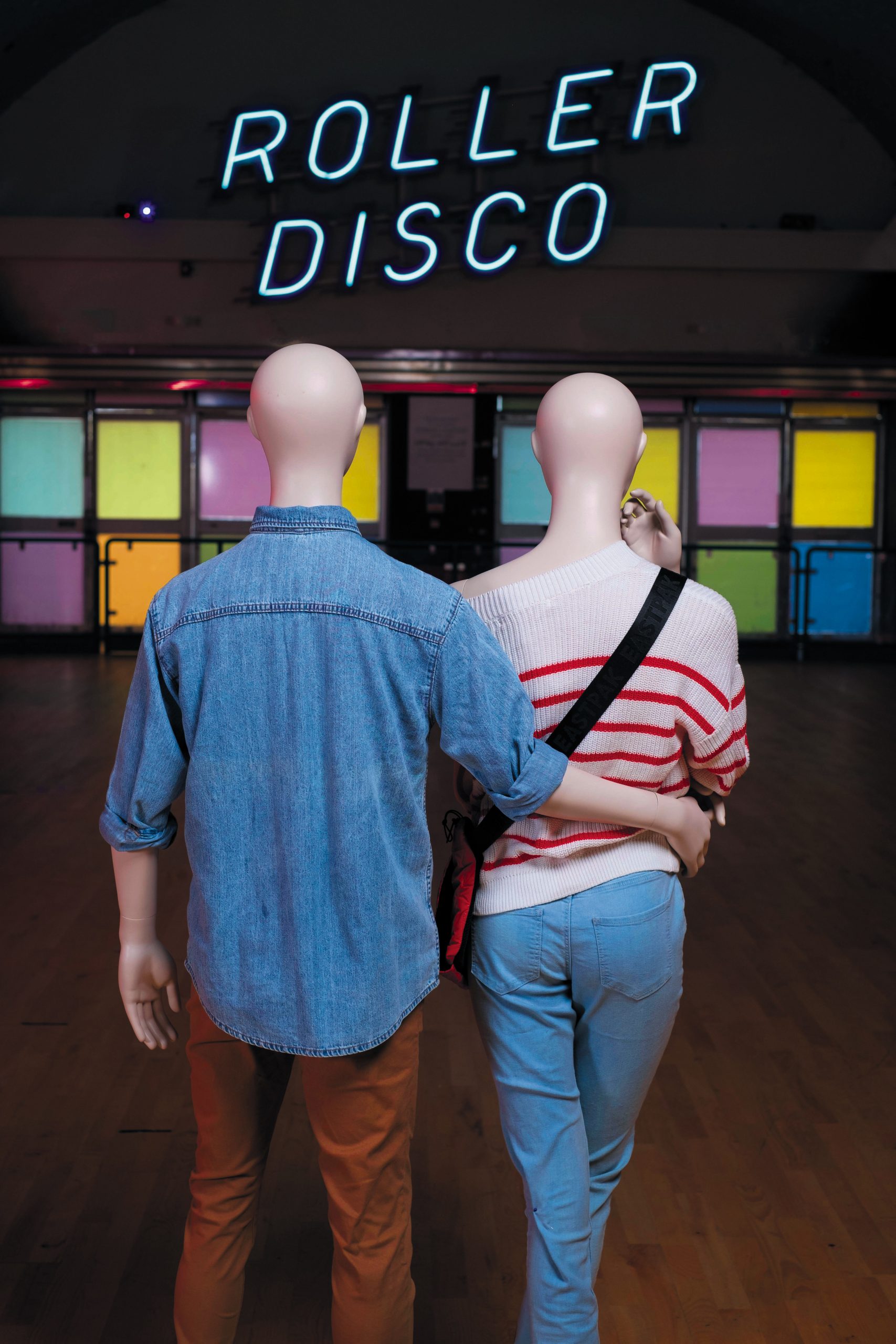 Mannequin by HOL Two mannequins, one dressed in a denim shirt and brown pants and the other in a striped sweater and jeans, are walking arm-in-arm on a wooden floor under a neon sign that reads "Roller Disco." Colorful lit panels line the wall behind them, reminiscent of the vibrant Farringdon Youth scene.