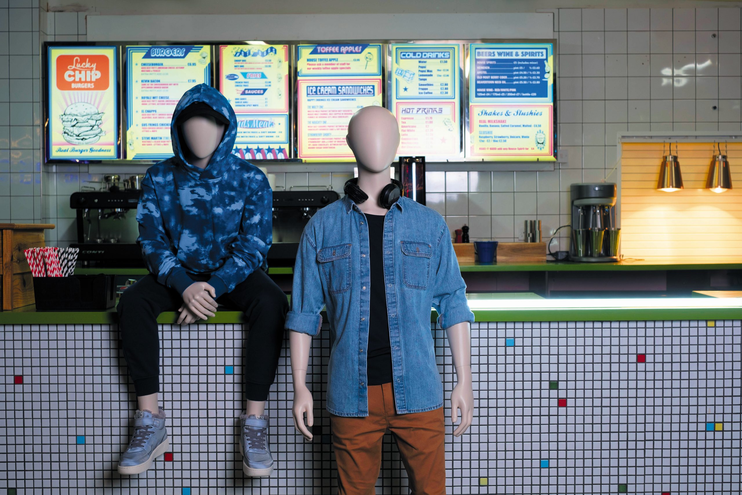 Mannequin by HOL Two mannequins in a retro-style cafe, one sitting on the counter wearing a blue hoodie and black pants, the other standing with headphones around its neck wearing a denim jacket and brown pants. A colorful menu board hangs in the background above a tiled counter, capturing that distinct Farringdon Youth vibe.