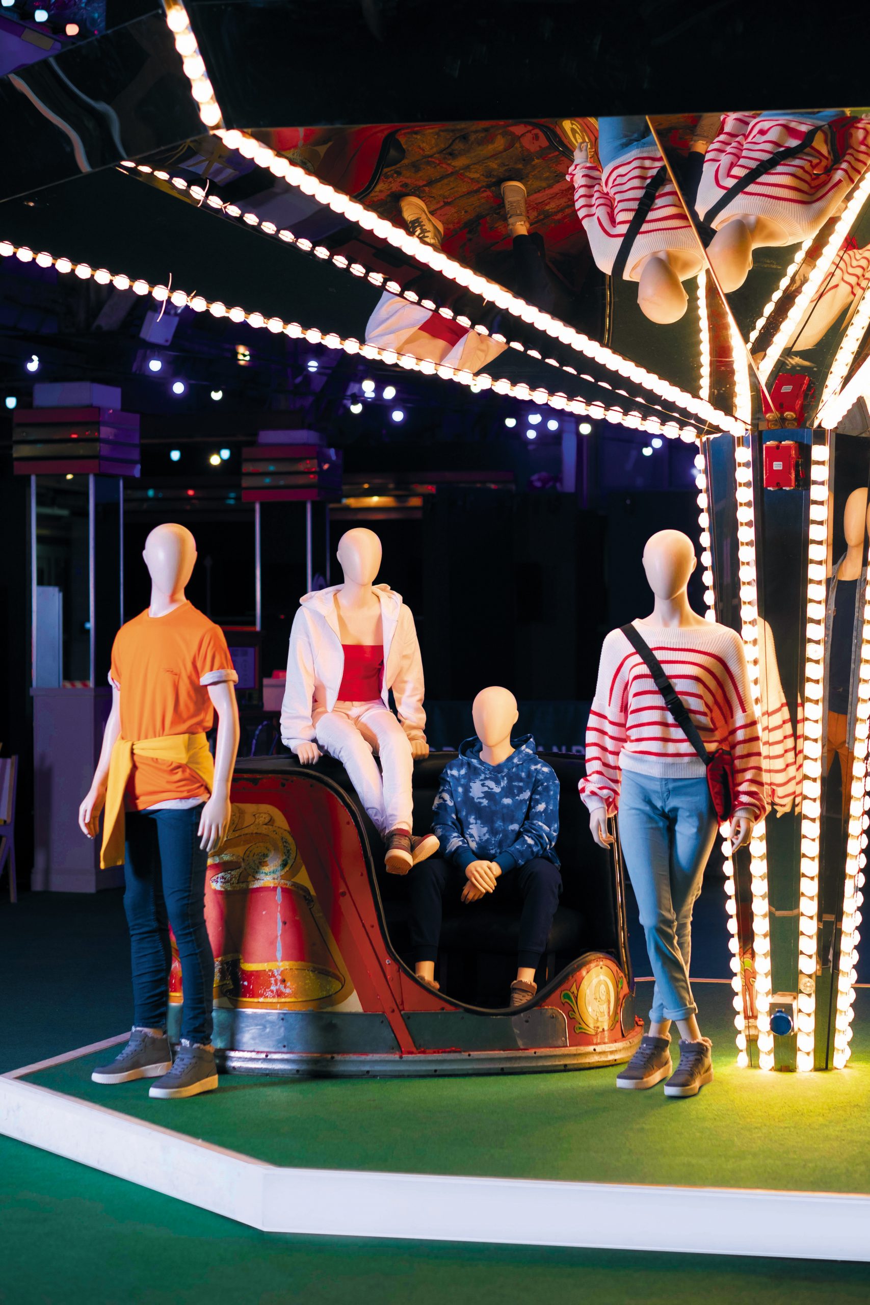Mannequin by HOL A group of mannequins dressed in casual clothing, including jeans, jackets, and tops, is displayed in a colorful, lit-up environment reminiscent of Farringdon. One mannequin is seated in a carnival ride, while others stand nearby. The setting features vibrant lights and a festive atmosphere buzzing with youthful energy.