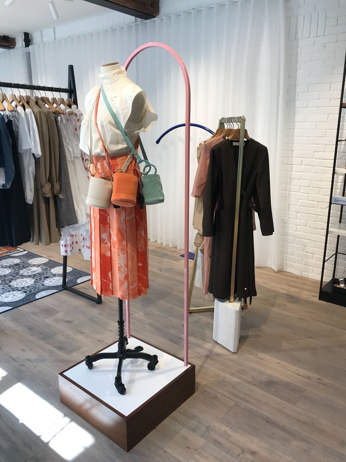Mannequin by HOL A retail display featuring a mannequin dressed in white and orange clothing with multiple crossbody bags in different colors. The display is set against a backdrop of hanging garments in a modern, well-lit store with white brick walls and wooden flooring.