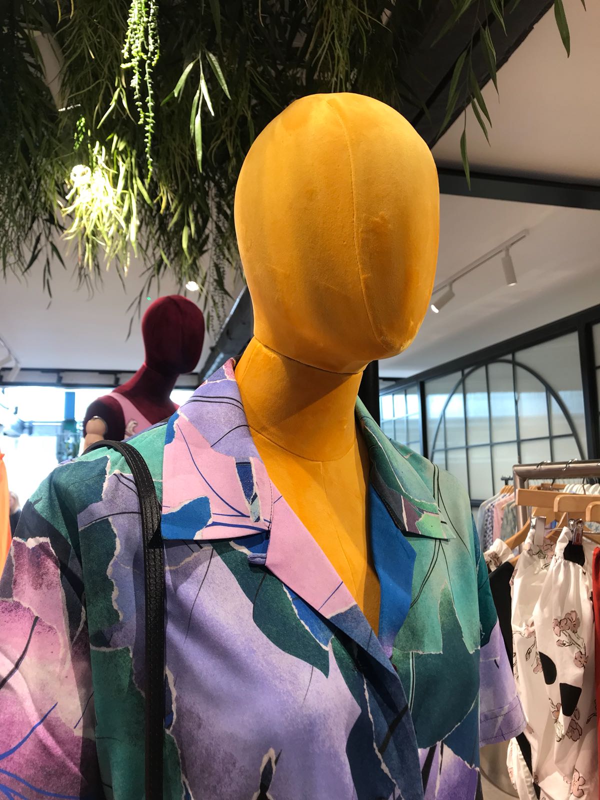 Mannequin by HOL A brightly colored mannequin with a smooth, yellow head and no facial features stands in a clothing store. It is dressed in a multicolored shirt with abstract patterns. The store has modern decor, with hanging plants visible at the top of the image.