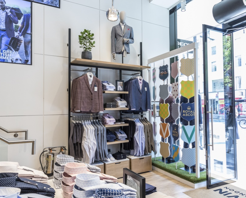 Mannequin by HOL A well-lit clothing store interior featuring neatly arranged men's suits on mannequins and hangers. There is a shelf with folded shirts, ties, and accessories. To the right, a patterned wall and an open glass door provide a view of the street outside.