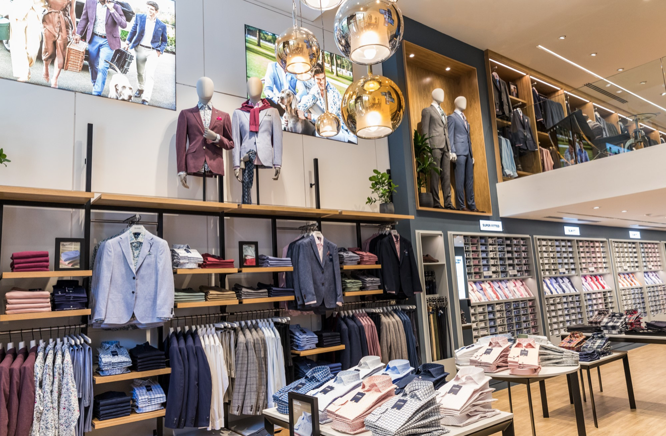 Mannequin by HOL A well-organized clothing store displays men's fashion items. Suits and blazers are on racks and mannequins, with folded shirts and accessories on tables. The store has modern lighting fixtures and neatly arranged shelves with shirts. Two large screens show fashion images.