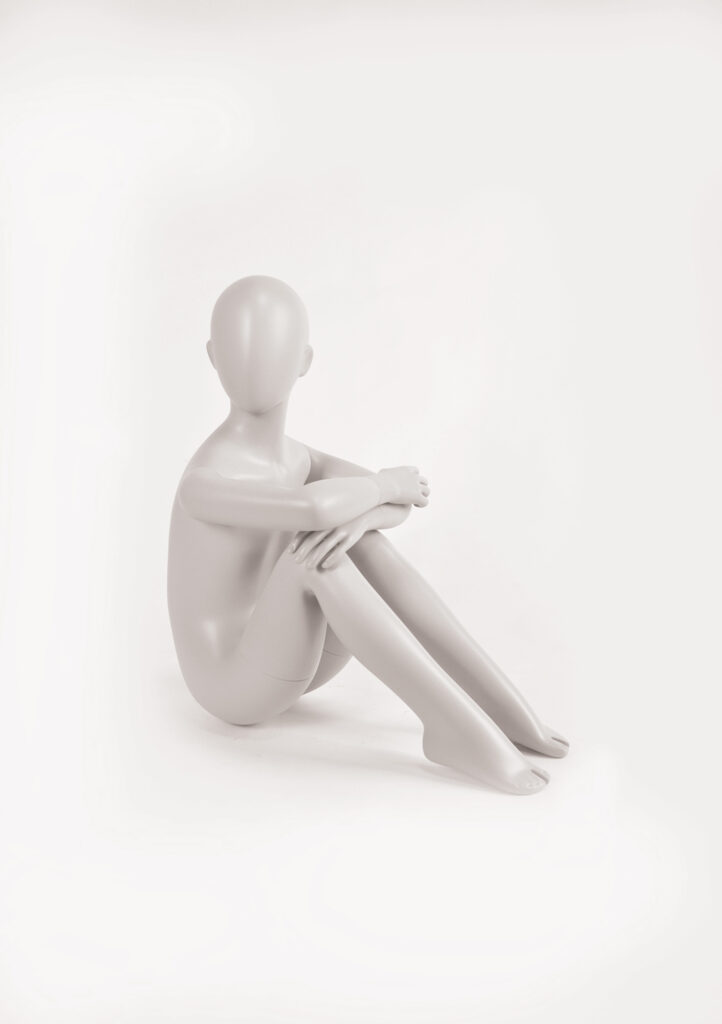 Mannequin by HOL A smooth, featureless, grey mannequin is seated on the ground with arms wrapped around its knees and legs bent. The background is plain and white, emphasizing the simple, abstract form of the FK05 mannequin from Farringdon Kids.