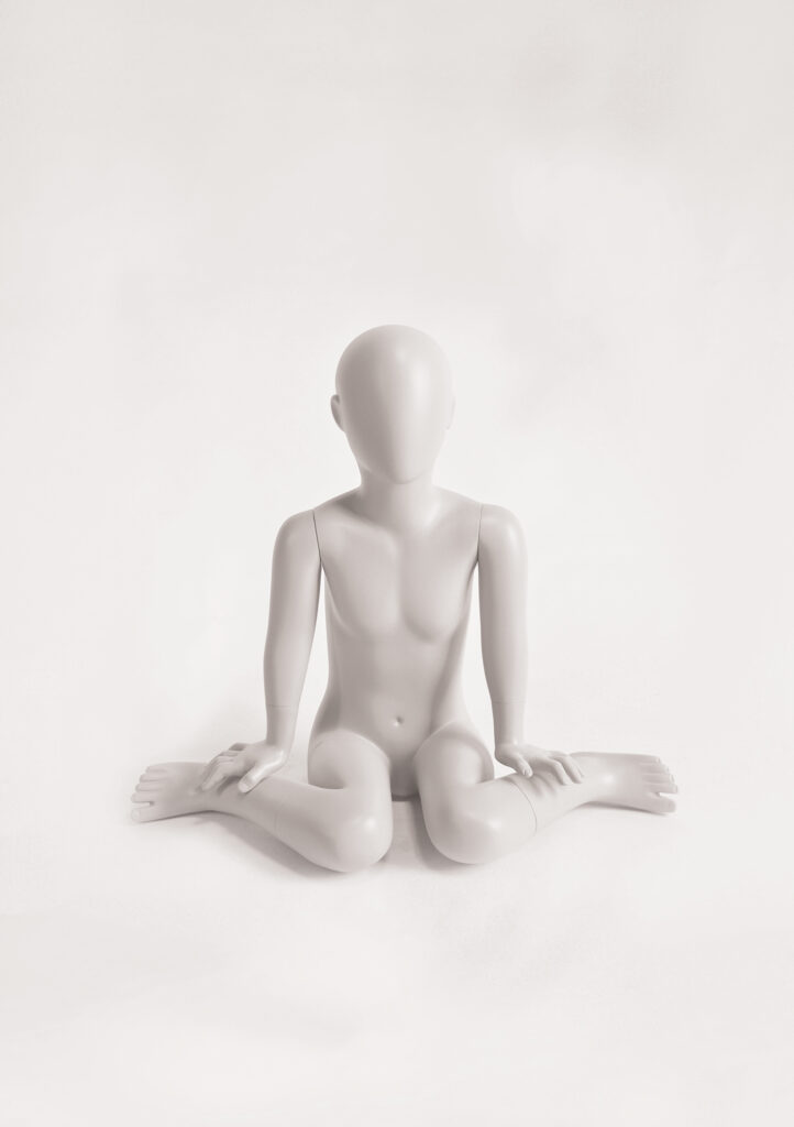 Mannequin by HOL A gray, featureless mannequin, reminiscent of the FK06 collection, is sitting on the floor with legs crossed and hands resting on the ground. The plain white background emphasizes the simplicity of its form, much like a Farringdon Kids display piece.