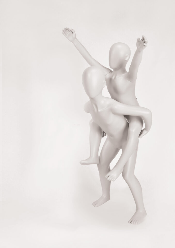 Mannequin by HOL Two faceless, featureless mannequins are engaged in a playful pose. One mannequin is piggybacking on the other, their arms extended upward and outward against a plain white background, suggesting movement and joy reminiscent of the carefree spirit of FK10 from Farringdon Kids.